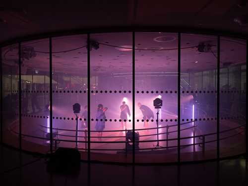 Fire mennesker danser inne i et glassrom lyst opp av lyskastere med lilla lys. Fotografi