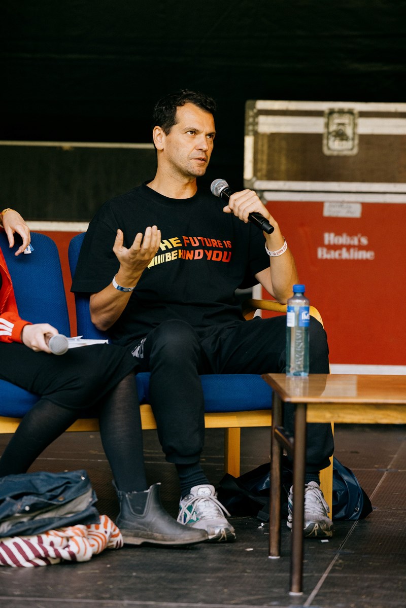 Performancekunstner Manuel Pelmus sitter på scenen og snakker i en håndholdt mikrofon. Fotografi