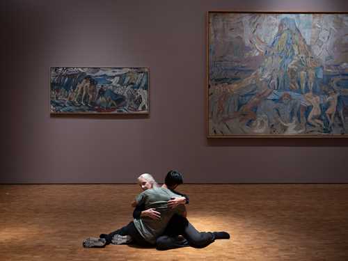 To mennesker sitter på gulvet i en omfavnelse. I bakgrunnen ser du maleriet Menneskeberget av Edvard Munch. Fotografi