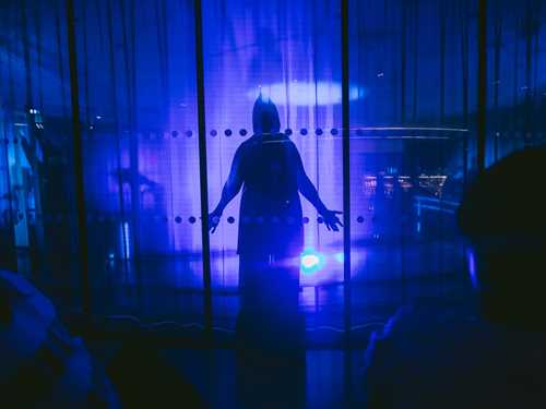En kappekledd skikkelse står bak en glassvegg i et rom lyst opp av blått lys. Fotografi